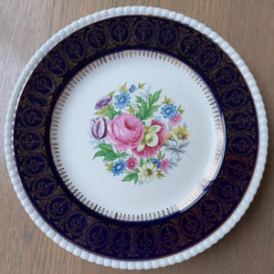May include: A white ceramic dinner plate with a blue and gold rim. The center of the plate features a floral design with pink, yellow, and blue flowers.