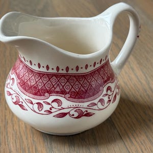 May include: Cream-colored ceramic creamer with a red floral and geometric pattern. The creamer has a curved spout and a handle. The design includes diamond shapes, dots, and leaf-like motifs. A classic piece for serving cream or milk.
