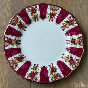 May include: A round white porcelain plate featuring a scalloped edge with a gold-tone rim. The border is decorated with alternating panels of deep magenta and floral clusters of pink and yellow roses tied with pink ribbons in a radial pattern.