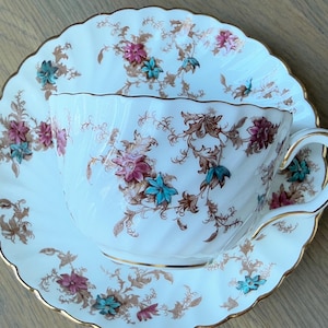 May include: A white teacup and saucer with a floral pattern in shades of brown, pink, and blue. The cup and saucer have a gold trim.