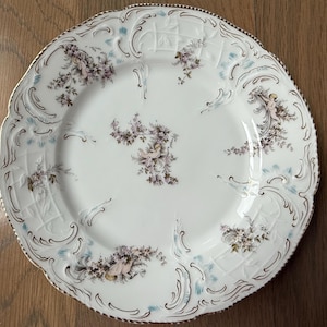 May include: A white porcelain plate with a gold rim and intricate floral designs. The plate features delicate blue and pink flowers and cherubs.