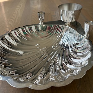 May include: A polished silver-toned serving dish shaped like a large scallop shell. The dish features a wide, fluted basin and is accented with three small, upright cylindrical containers attached to the back edge, including one larger central cup.