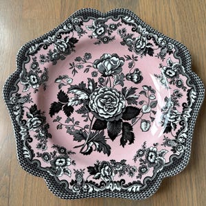 May include: A decorative pink plate with a floral design. The plate features a central arrangement of large black and white roses surrounded by smaller floral motifs. The scalloped edge is detailed with a black and white checkered pattern.