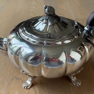 May include: A silver teapot with a rounded, bulbous body and a wooden handle. The teapot has a decorative lid with a finial and sits on three feet.