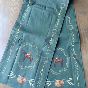 May include: A dark green table runner featuring a repeating equestrian pattern. The design includes illustrations of a rider on a brown horse framed by a laurel wreath, brown saddles, gold stirrups, and gold bits, accented by light green leaf sprigs.