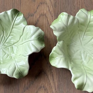 May include: Two light green ceramic leaf-shaped dishes. The dishes have a detailed vein pattern and a glossy finish. The color is a soft, muted green, with darker green accents. The dishes are placed on a wooden surface.