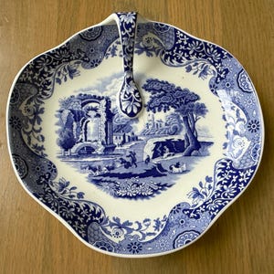 May include: A blue and white ceramic serving dish with a handle. The dish features a pastoral scene in the center and a floral border. The handle is also decorated with floral patterns. The dish is on a wooden surface.