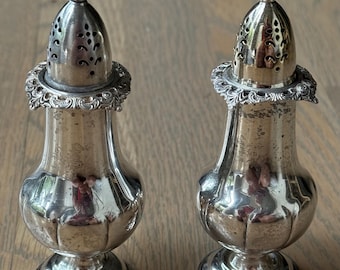 May include: Two antique silver salt shakers. Each shaker has a bulbous body, a decorative filigree collar, and a perforated top. The shakers are highly reflective, with a small finial on top. The salt shakers are displayed on a wooden surface.