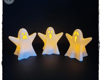 May include: Three white ghost-shaped candle holders with yellow glowing faces. The ghosts are standing on a dark surface.
