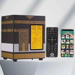 May include: A black and gold electronic clock in the shape of the Kaaba, with a digital display showing the time, date, and temperature. A remote control and a smartphone are next to the clock. The smartphone displays images of religious figures.