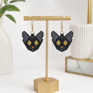 Peut inclure: Une paire de boucles d'oreilles pendantes représentant une tête de chat en perles. Les visages sont composés de petites perles noires, grises et jaunes, avec des yeux jaunes vifs et un petit nez orange. Elles sont suspendues à des crochets dorés.
