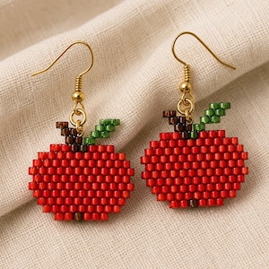 May include: A pair of dangle earrings featuring apple-shaped charms crafted from small red, green, and brown beads. The apples have a round red body, a small brown stem, and a single green leaf. Each charm hangs from a gold-toned metal hook.