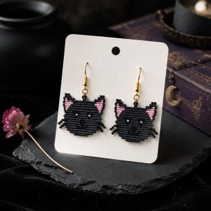 May include: A pair of beaded earrings shaped like cat heads, crafted from small black beads with pink accents on the ears and white bead details for the eyes and nose. The earrings feature gold-toned metal fishhook wires for attachment.