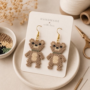 May include: Handmade beaded teddy bear earrings with gold-coloured hooks. The earrings are tan, cream, and black, with a green collar. The earrings are displayed on a white card that reads "HANDMADE with love."
