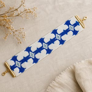 May include: A wide beaded bracelet featuring a geometric pattern in royal blue, white, and light blue. The bracelet is finished with gold-tone metal end bars and a lobster claw clasp, creating a structured band with a repeating cross motif.