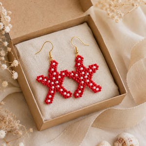 May include: A pair of dangle earrings shaped like starfish, crafted from small red and white beads. The earrings feature gold-toned metal hooks for attachment and are presented inside a square cardboard box lined with light beige fabric.