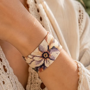 Pode incluir: Uma pulseira larga de contas com um design floral. A pulseira apresenta uma flor grande com pétalas creme, roxas e castanhas, centrada com um círculo castanho escuro. A pulseira é usada no pulso.