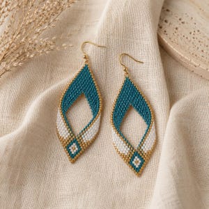May include: A pair of teardrop earrings with a beaded design. The earrings have a teal and white pattern, edged with gold beads. They hang from gold-coloured ear wires, and are displayed on a beige fabric.