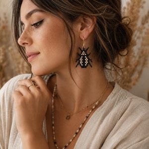 May include: Black and gold beaded bug earrings with gold-colored hooks. The earrings are a pixelated design, with a black body and gold-colored accents. The earrings are worn by a person with a light-colored top and multiple necklaces.