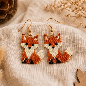 May include: A pair of beaded fox earrings. Each earring features a fox design with orange, white, black, and gold beads. The earrings have gold-colored ear wires and are displayed on a beige fabric surface.