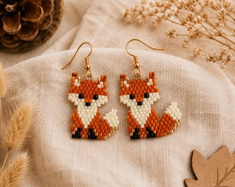 May include: A pair of dangle earrings featuring a fox design crafted from small orange, white, and black beads. The fox figures are suspended from gold-toned metal hooks. Each earring depicts a sitting fox with a bushy tail and pointed ears.