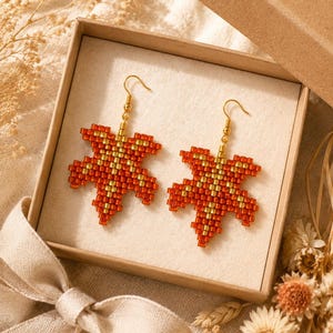 May include: A pair of dangle earrings crafted from small orange and gold beads in the shape of maple leaves. Each earring features a gold-toned metal hook for attachment, showcasing a detailed geometric pattern created by the arrangement of the beads.
