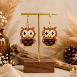 May include: A pair of owl-shaped dangle earrings crafted from small brown, dark brown, white, and yellow beads. The earrings feature gold-toned metal hooks and are displayed on a gold-toned T-shaped stand with a rectangular wooden base.