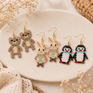 Peut inclure: Trois paires de boucles d'oreilles pendantes en perles représentant des animaux. Une paire montre des ours bruns au ventre crème, une paire des lapins aux tons pastel, et une paire des pingouins noirs, blancs et rouges. Crochets dorés.