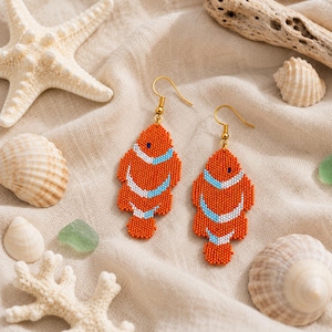 May include: A pair of dangle earrings crafted from small orange, white, and light blue beads in the shape of clownfish. Each earring features a gold-toned metal hook for attachment, showcasing a detailed beaded pattern that mimics the fish's body.