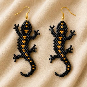 May include: A pair of drop earrings shaped like salamanders, crafted from small black and gold beads. Each earring features a gold-toned metal hook for attachment, with the beaded lizard design displaying a black body and a central gold stripe.