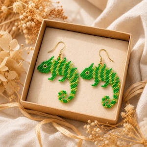 Puede incluir: Un par de pendientes colgantes con un diseño de camaleón hecho de cuentas en tonos verde brillante y verde lima. Cada camaleón tiene una cola curva y un pequeño ojo negro, suspendido de ganchos de metal dorado dentro de una caja cuadrada.