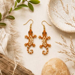 May include: A pair of drop earrings featuring beaded lizard charms in shades of orange, white, and brown. The lizards have small black eyes and are attached to gold-toned metal fishhook ear wires. The beaded bodies display a detailed pattern.