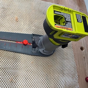 May include: A green and gray cordless router with a black and gray base plate, sitting on a white mat. The router has a battery pack with a warning label that reads "WARNING:  A POTENTIAL HAZARD".