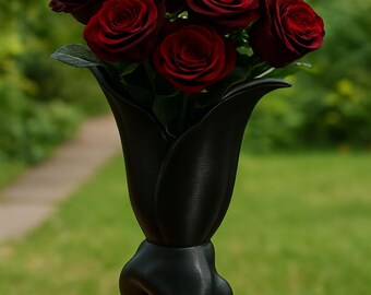 May include: A display of dark red roses in a black, artistic vase. The vase features an unusual, flowing design with a wide top and a base shaped like stylized legs. The roses are fully open, providing a vivid contrast against the dark vase.
