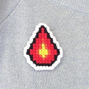 Könnte beinhalten: Ein tropfenförmiger Kreuzstich-Aufnäher mit einem verpixelten Flammendesign in Rot-, Orange- und Gelbtönen, schwarz umrandet und mit einer dicken weißen Häkelkante versehen, befestigt auf einem hellblauen Strickpullover mit silbernen Verzierungen.