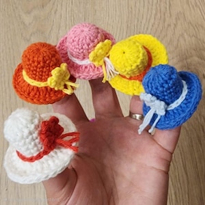 Może przedstawiać: Dłoń prezentuje pięć miniaturowych kapeluszy zrobionych na szydełku w różnych kolorach: pomarańczowym, różowym, żółtym, niebieskim i białym. Każdy kapelusz ma kontrastującą wstążkę lub kwiat. Kapelusze są małe, prawdopodobnie dla lalek lub rękodzieła.
