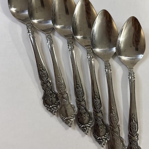 Puede incluir: Un juego de siete cucharas de color plateado. Cada cuchara tiene un cuenco redondeado y un mango decorativo con detalles florales y de volutas. Las cucharas están dispuestas en diagonal sobre una superficie blanca.