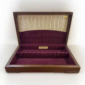 May include: Open wooden silverware storage box with a dark brown exterior and a deep burgundy velvet interior. The box has multiple compartments for utensils and a cream-colored fabric lining the lid. A small gold-colored label is visible.