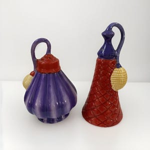 May include: A pair of ceramic salt and pepper shakers. One is purple with a red top and a gold accent. The other is red with a blue top and a gold accent. Both have a handle and a decorative design.