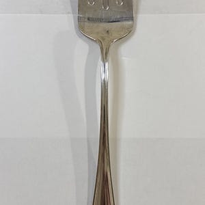 Può includere: Una forchetta da portata argentata con quattro rebbi e un lungo manico affusolato. Il manico ha un motivo floreale decorativo alla base. La forchetta è progettata per servire il cibo.