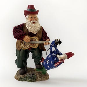 Puede incluir: Una figura de Papá Noel vestido con un sombrero de vaquero y una camisa a cuadros, tocando la guitarra. Se encuentra sobre una pequeña base texturizada. En la otra mano sostiene un saco azul con copos de nieve blancos y rayas rojas y blancas.