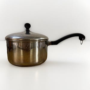May include: A stainless steel saucepan with a lid and a black handle. The pot has a shiny, reflective surface and a black knob on the lid. The handle curves out and has a small hole at the end. Kitchenware.