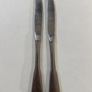 May include: Two stainless steel butter knives. The blades are curved, and the handles have a slender, elongated shape. The knives are set against a plain, light-coloured background. The knives are suitable for spreading butter.