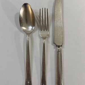 May include: Silverware set including a spoon, fork, and knife. The spoon has an oval bowl, the fork has four tines, and the knife has a rounded blade. Each piece has a decorative design at the base of the handle.