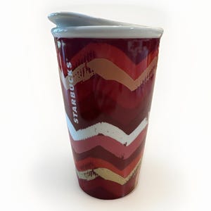 May include: A red Starbucks travel mug with a white lid. The mug features a chevron pattern in shades of red, white, and gold. The Starbucks logo is printed vertically on the side of the mug. The mug is designed to keep beverages hot or cold.