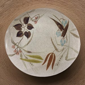 May include: Round ceramic plate with a speckled off-white background. The plate features a floral design with brown leaves, pink berries, and blue bell-shaped flowers. The plate has a glossy finish and is suitable for serving food.