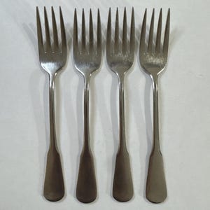 May include: Four silver forks with tines and long handles. The forks are arranged side by side, with the tines pointing upwards. The handles have a rounded, slightly flattened shape. The forks are made of a shiny, reflective metal.