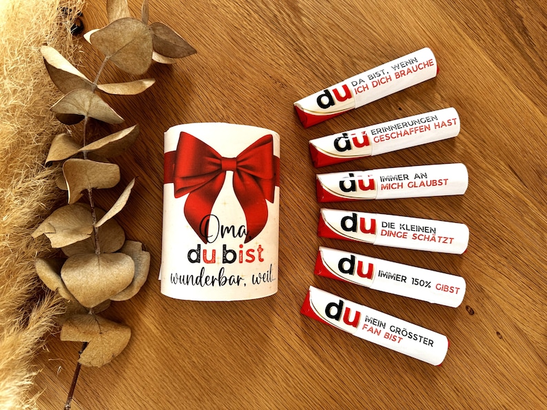 May include: A white gift box with a red bow and the text "Oma du bist wunderbar, weil" on it.  There are five white tubes with red and gold accents and the word "du" on them. Each tube has a different message on it.