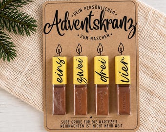 May include: A rectangular brown cardboard display card featuring the text "DEIN PERSÖNLICHER Adventskranz ZUM NASCHEN" and "SÜBE GRÜBE FÜR DIE WARTEZEIT – WEIHNACHTEN IST NICHT MEHR WEIT." Four chocolate bars are attached, each with a yellow candle-shaped label.