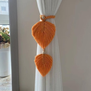 May include: Orange macrame leaf curtain tiebacks with wooden beads. The tiebacks are attached to a sheer white curtain. The leaves are a vibrant orange color and have a textured, fringed appearance. The curtain tiebacks are a decorative home decor item.